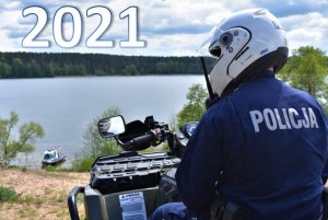 Policjant na kładzie, w tle jezioro. Na zdjęciu napis 2021.
