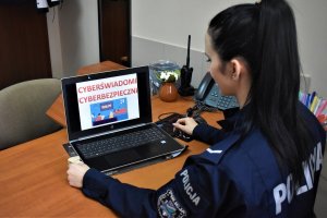 Policjantka siedzi przy biurku. Na monitorze napis cyberświadomi, cyberbezpieczni.