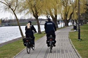 Policjanci jadą jednośladami po ścieżce rowerowej koło rzeki.