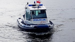policyjna łódź motorowodna, policjanci siedzą w motorówce jeden z nich trzyma ster, policjant stoi w łódce i obserwuje otoczenie, policjanci sprawdzają uprawnienia kierowcy łodzi, policjant zatrzymuje łódź do kontroli