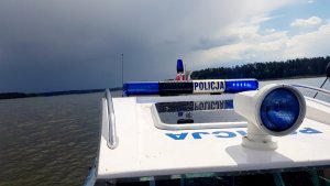 policyjna łódź motorowodna, policjanci siedzą w motorówce jeden z nich trzyma ster, policjant stoi w łódce i obserwuje otoczenie, policjanci sprawdzają uprawnienia kierowcy łodzi, policjant zatrzymuje łódź do kontroli