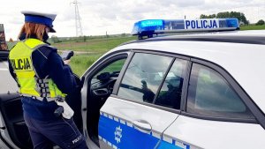 policjantka mierzy prędkość fotoradarem, policjantka siedzi w radiowozie i na urządzeniu mobilnym sprawdza tożsamość osoby kontrolowanej, stojący na poboczu radiowóz,  stojąca przy radiowozie policjantka