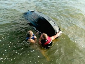 dwie osoby w wodzie trzymają się skutera wywróconego do góry dnem
uratowane osoby i policjant na łodzi policyjnej