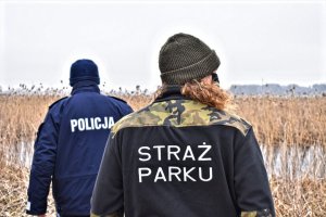 Policjant i funkcjonariuszy straży parku stoją tyłem. W tle rzeka Biebrza.