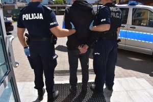 policjantka zakłada kajdanki osobie zatrzymanej, na ręce trzymane z tyłu. mężczyzna prowadzony przez dwóch policjantów. mężczyzna trzymany za ręce przez dwóch policjantów, stoją na schodach przed budynkiem