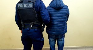 umundurowany policjant trzyma za rękę w zgięciu łokciowym zatrzymanego ubranego w niebieską kurtkę