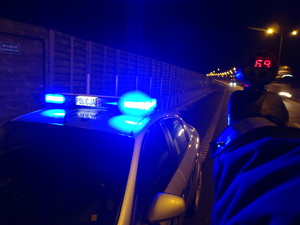 noc ulica, oznakowany radiowóz na sygnałach obok radiowozu stoi policjant i mierzy prędkość radarem nadjeżdżających pojazdów