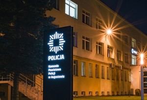 Zdjęcie obszaru przed komendą. Baner z napisem Komenda Powiatowa Policji w Augustowie.